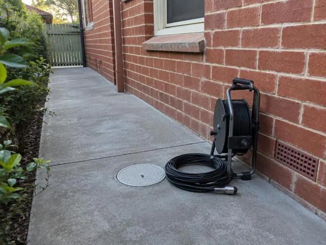 Brick house side path with a capped round drain access cover and a coiled drain camera reel beside it in Naremburn